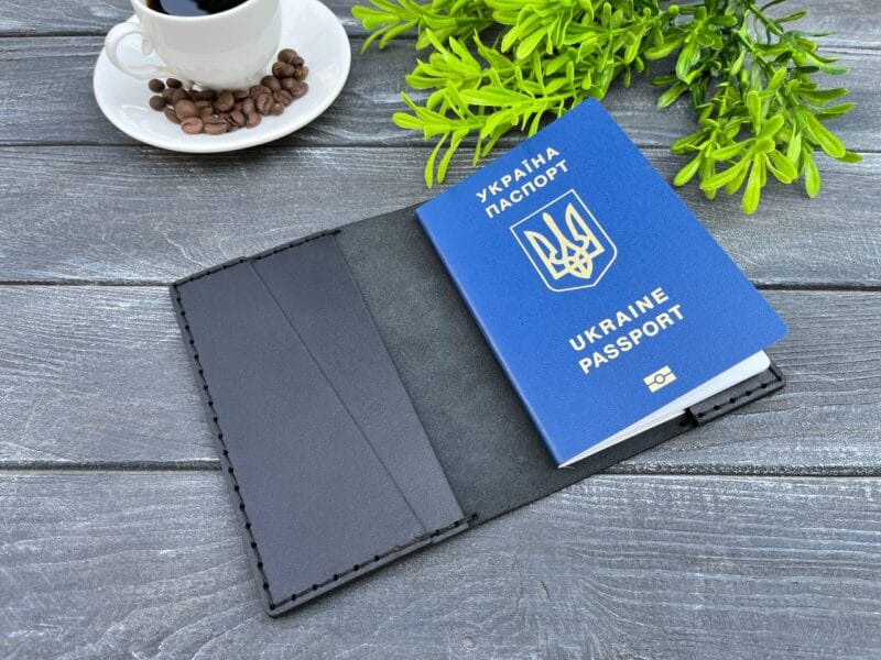 Відкрита чорна шкіряна обкладинка для паспорта, в якій лежить синій паспорт України. Обкладинка виготовлена з якісної шкіри з акуратними швами, розташована на дерев'яному фоні поруч з декоративною рослиною та чашкою кави.