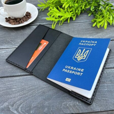Відкрита чорна шкіряна обкладинка для паспорта з вбудованим відділенням для банківської картки та українським біометричним паспортом всередині. Поруч на дерев'яній поверхні — декоративна зелень та чашка кави.