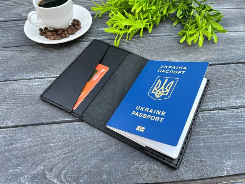 Відкрита чорна шкіряна обкладинка для паспорта з вбудованим відділенням для банківської картки та українським біометричним паспортом всередині. Поруч на дерев'яній поверхні — декоративна зелень та чашка кави.