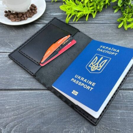 Відкрита чорна шкіряна обкладинка для паспорта, в якій знаходиться синій паспорт України та декілька пластикових карток. На задньому фоні чашка кави та зелена рослина.