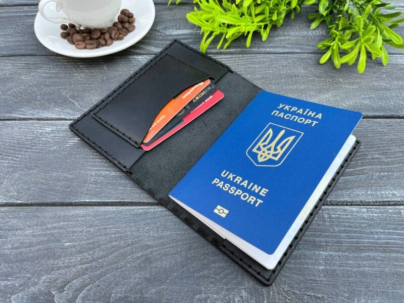 Відкрита чорна шкіряна обкладинка для паспорта, в якій знаходиться синій паспорт України та декілька пластикових карток. На задньому фоні чашка кави та зелена рослина.