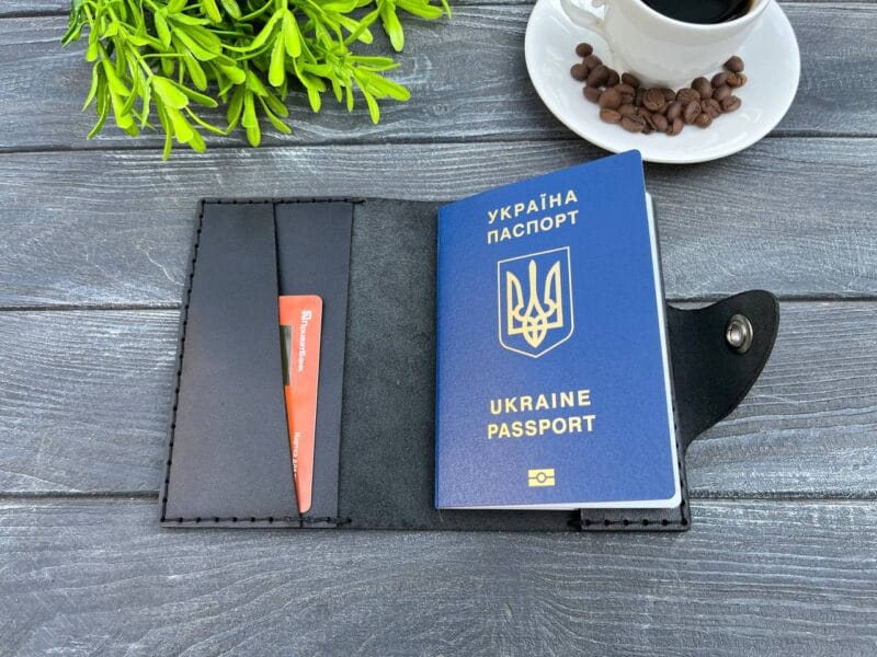 Відкрита чорна шкіряна обкладинка для паспорта з вставленим українським паспортом і кредитною карткою, розташована на дерев'яній текстурованій поверхні.