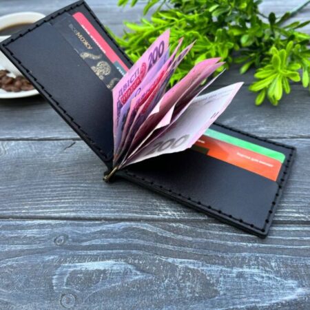 Відкритий чорний шкіряний затискач для купюр з купюрами і картками всередині, розміщений на дерев'яній поверхні поруч із чашкою кави.