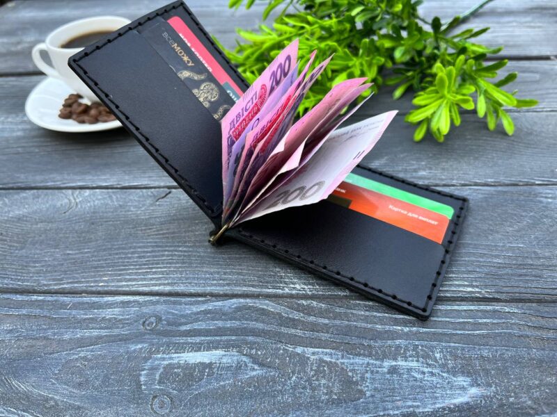 Відкритий чорний шкіряний затискач для купюр з купюрами і картками всередині, розміщений на дерев'яній поверхні поруч із чашкою кави.