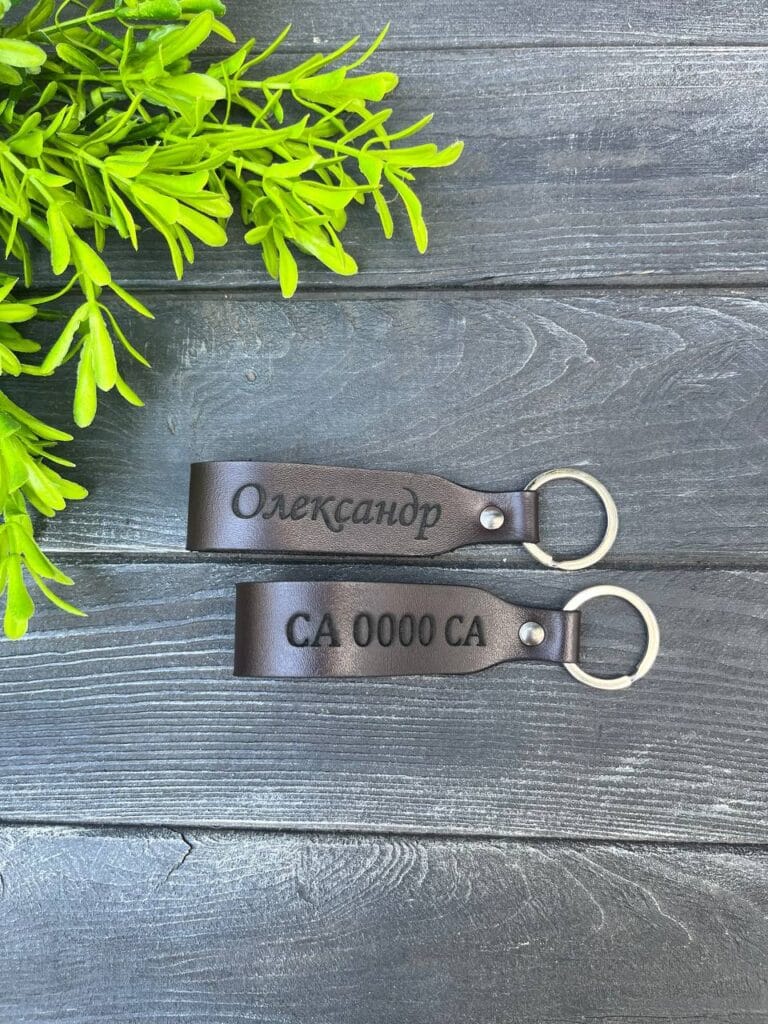 Два шкіряні брелоки з гравіруванням "Олександр" та "CA 0000 CA" на темно-сірому дерев'яному фоні.