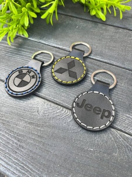 Три шкіряні брелоки з логотипами BMW, Mitsubishi та Jeep, розташовані на сірій дерев'яній поверхні.