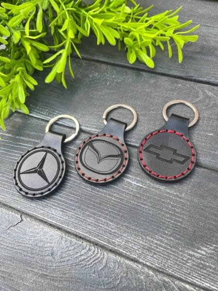 Шкіряні брелоки з логотипами Mercedes, Mazda та Chevrolet на темній поверхні з зеленою гілкою.