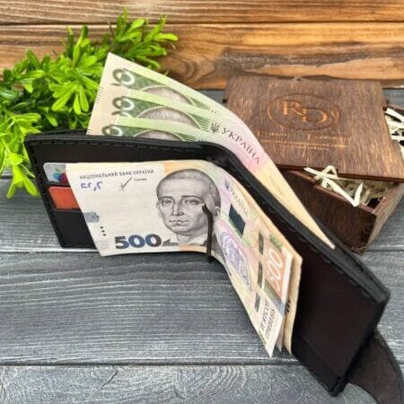 Чорний шкіряний гаманець, наповнений купюрами, розкритий на столі з дерев'яною текстурою, поруч декоративна зелень і дерев'яна коробка.