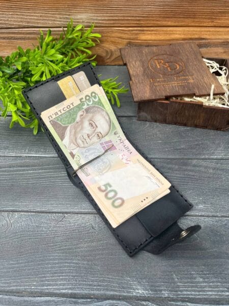 Відкритий чорний гаманець із купюрами номіналом 500 гривень, вкладеними у відділення для грошей, розташований на дерев'яній поверхні з декоративною зеленню та дерев'яною коробкою на задньому плані.